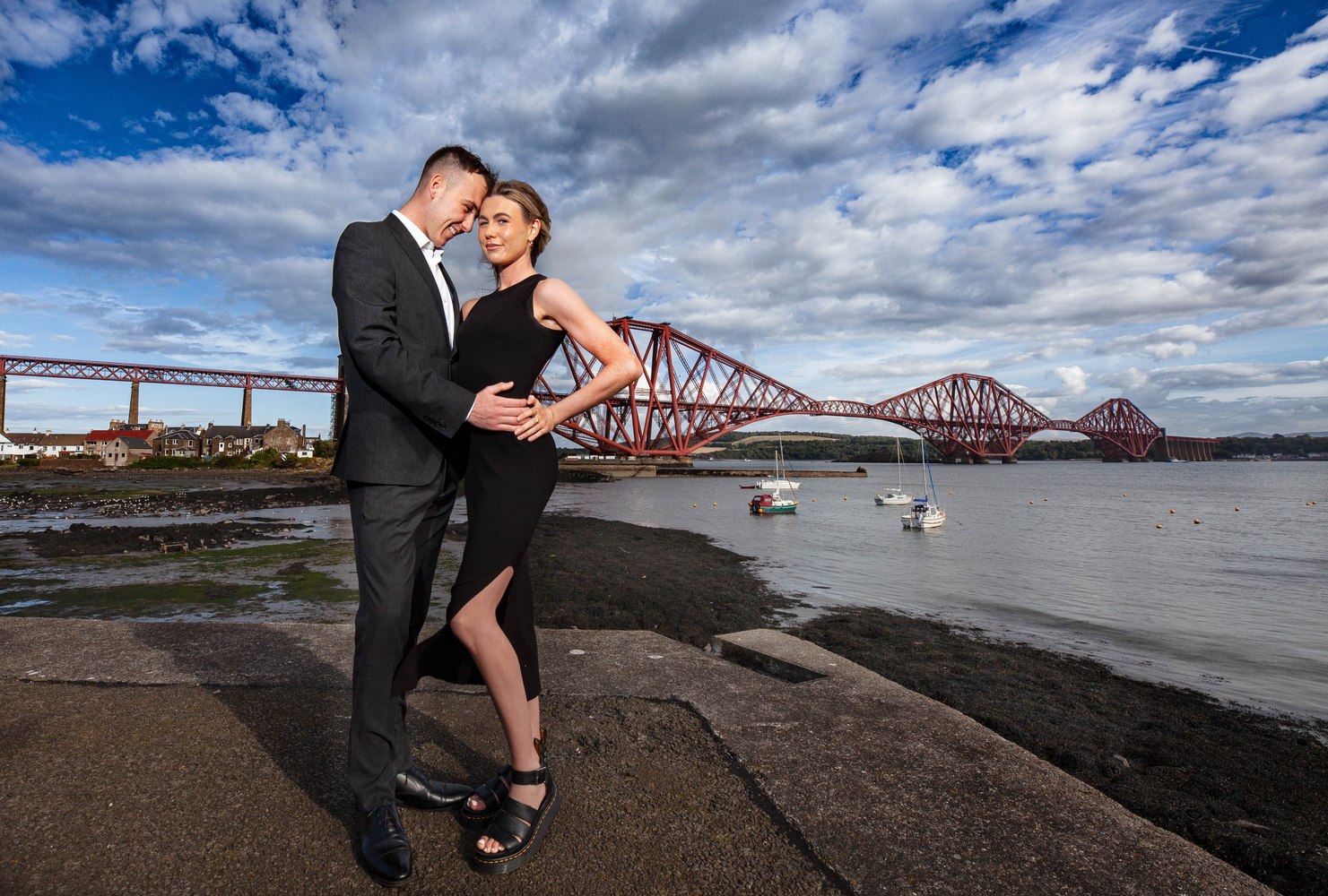 Couple embracing on the shoreline with a historic iron rail bridge behind them
