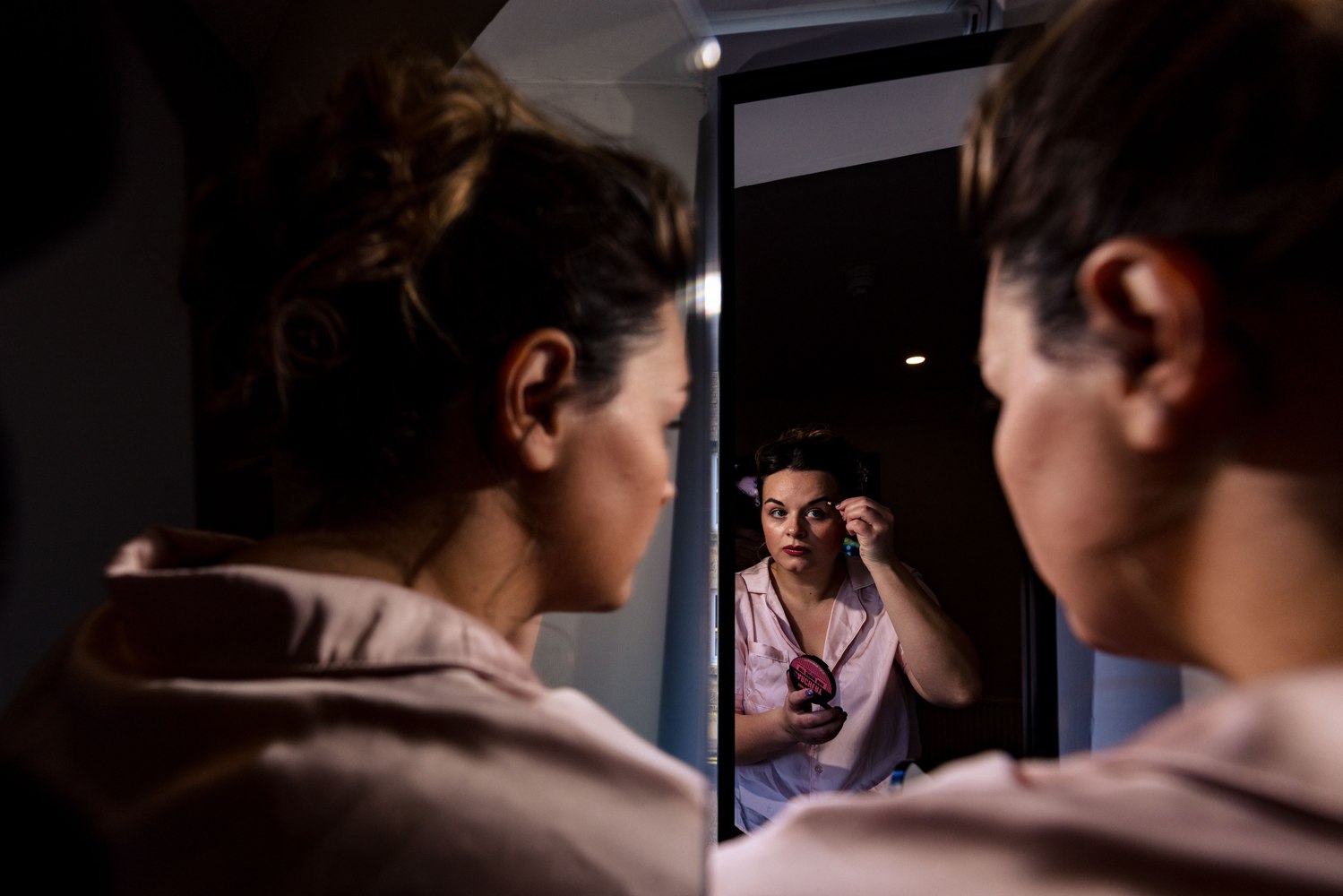 Bride applying makeup, reflected through an open doorway mirror