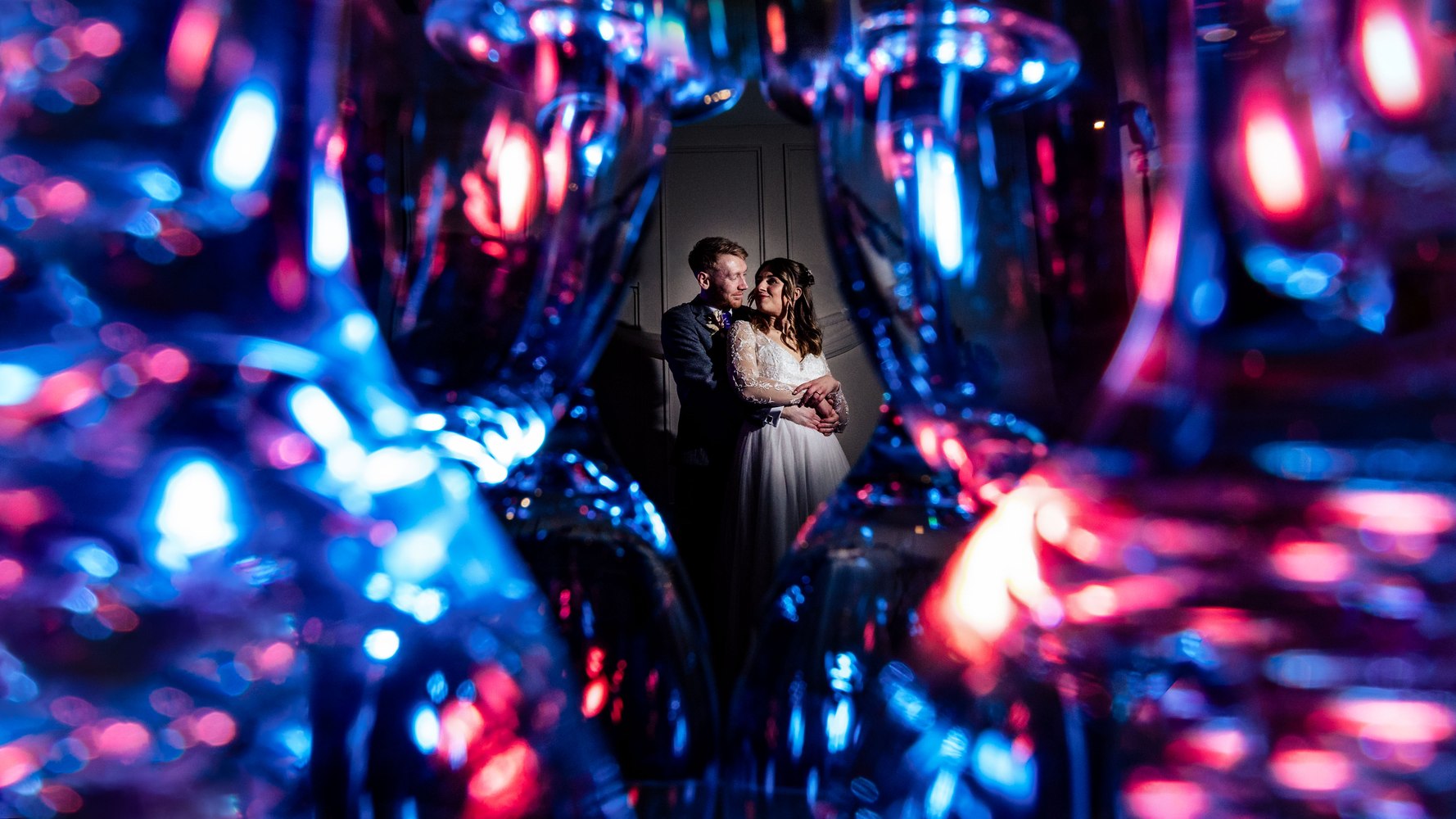 Couple embracing, framed through coloured glassware lit blue and pink