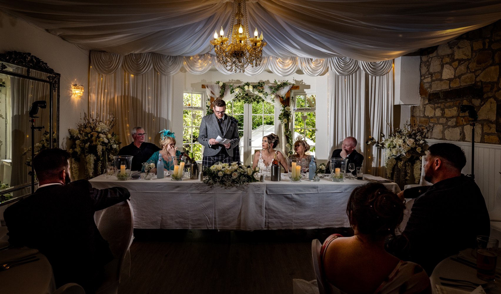 Groom delivering a speech to the room under a chandelier