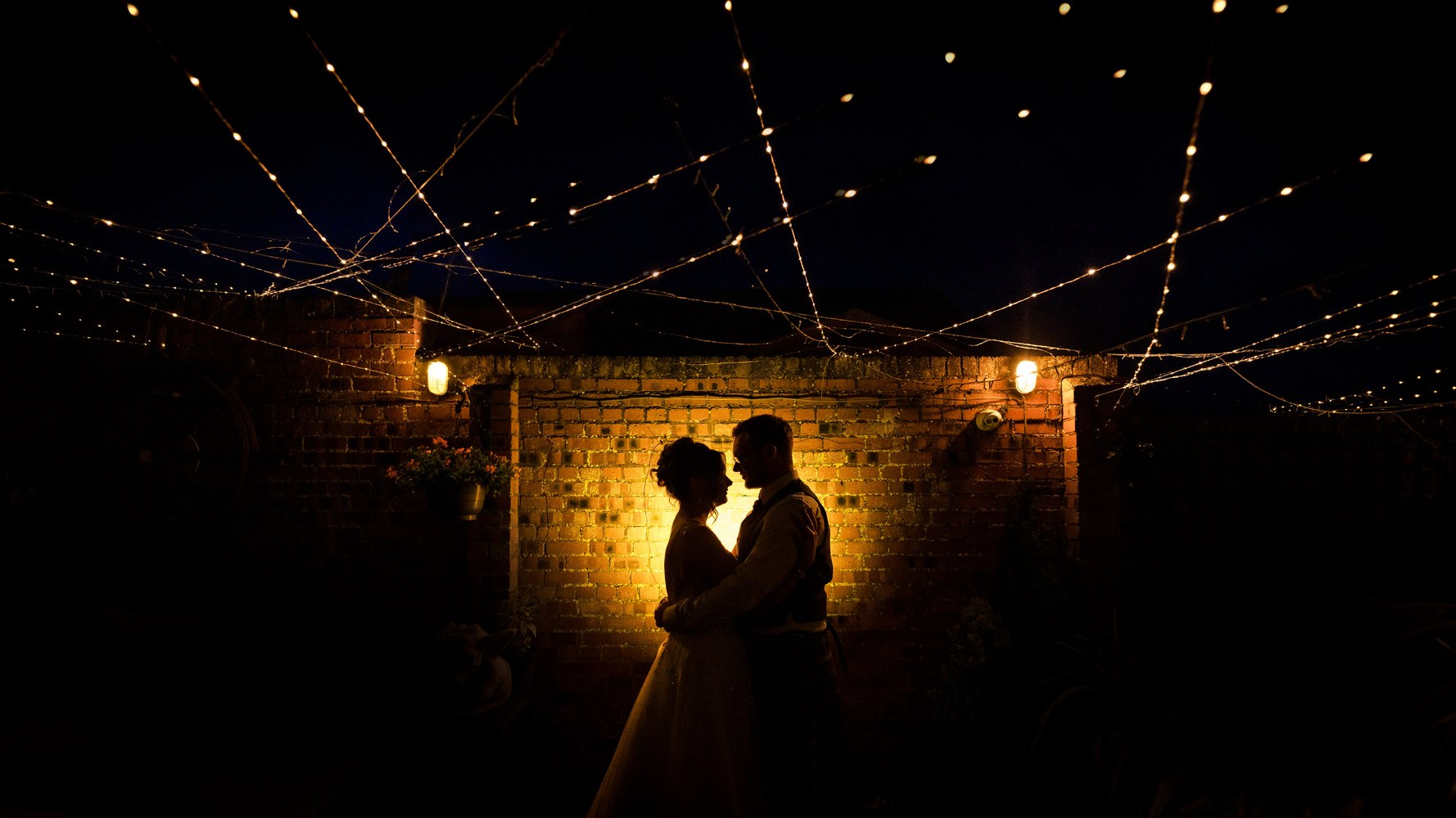 Couple silhouetted against string lights and a brick wall at night