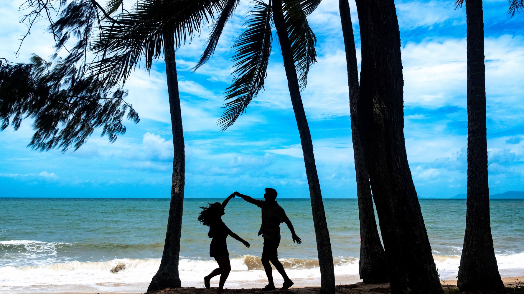 Couple dancing on the beach, silhouetted under palm trees