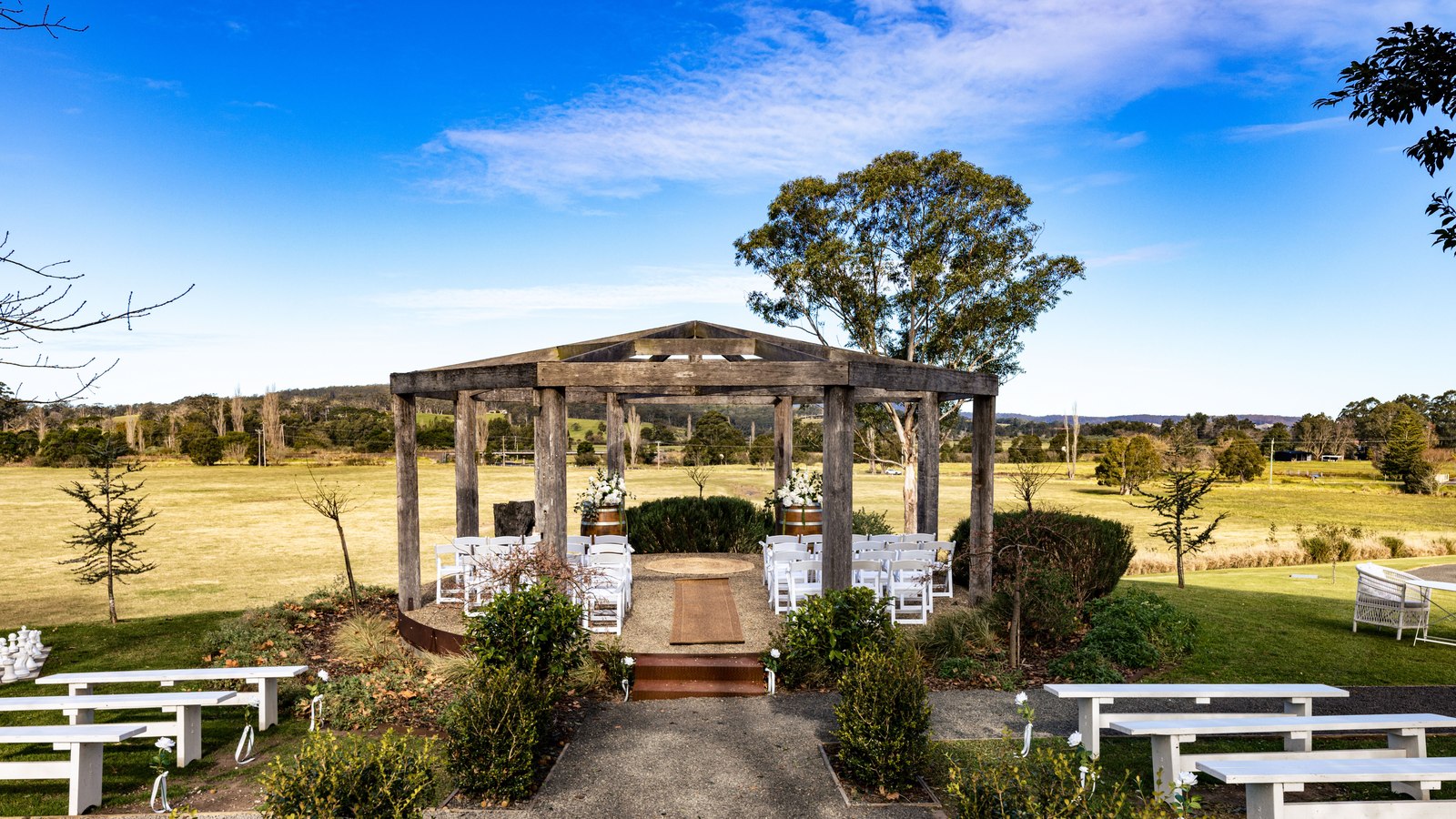 A rustic timber ceremony pergola set up with white chairs, overlooking open countryside